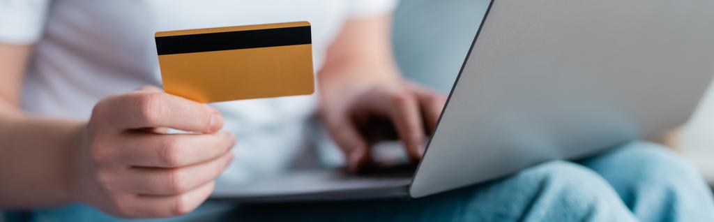 enfoque selectivo de la tarjeta de crédito en la mano de la mujer recortada con el ordenador portátil sobre fondo borroso, bandera - Foto, Imagen