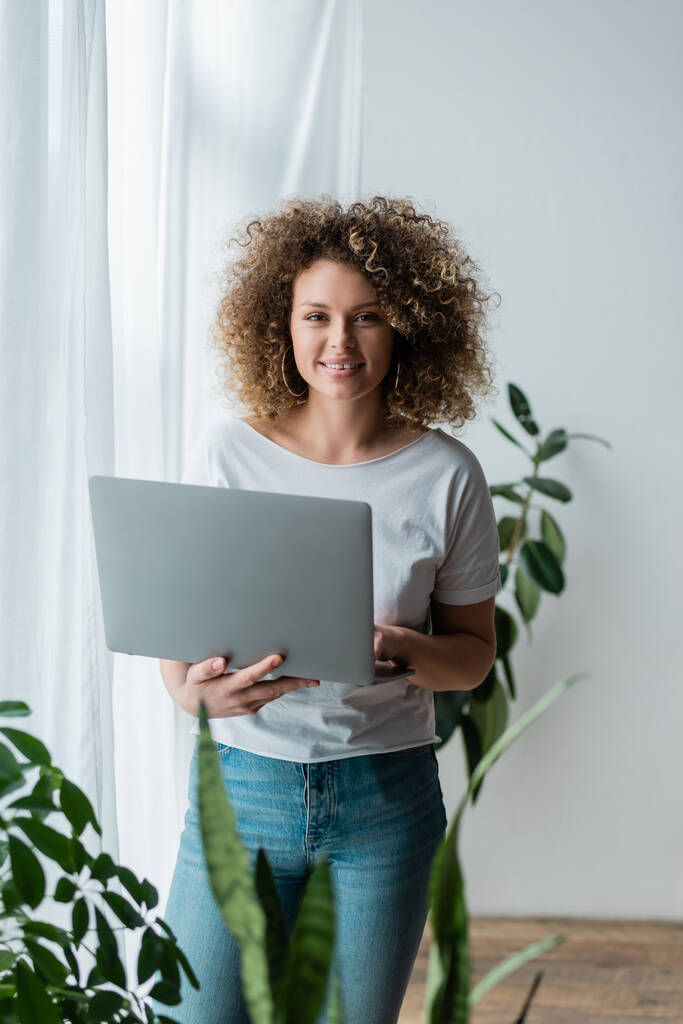 šťastná a kudrnatá žena stojící doma s laptopem a dívající se do kamery - Fotografie, Obrázek