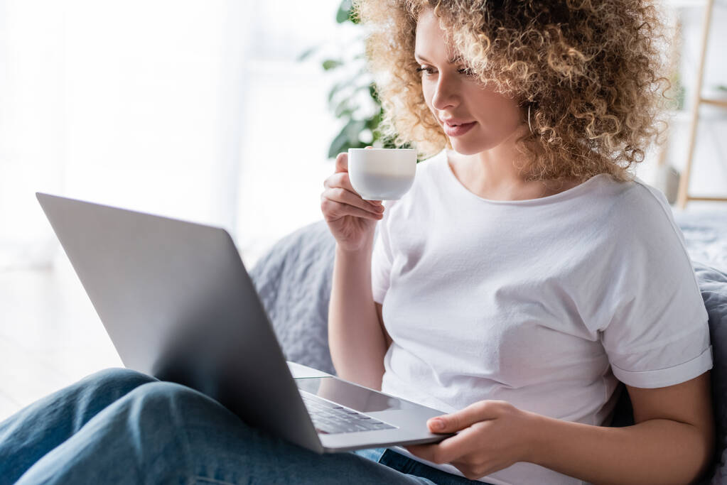 femme heureuse et bouclée avec tasse de café regardant ordinateur portable dans la chambre - Photo, image