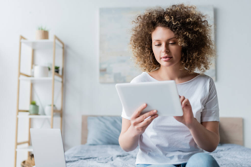 mujer con el pelo ondulado utilizando tableta digital mientras está sentado cerca de la computadora portátil en el dormitorio - Foto, Imagen