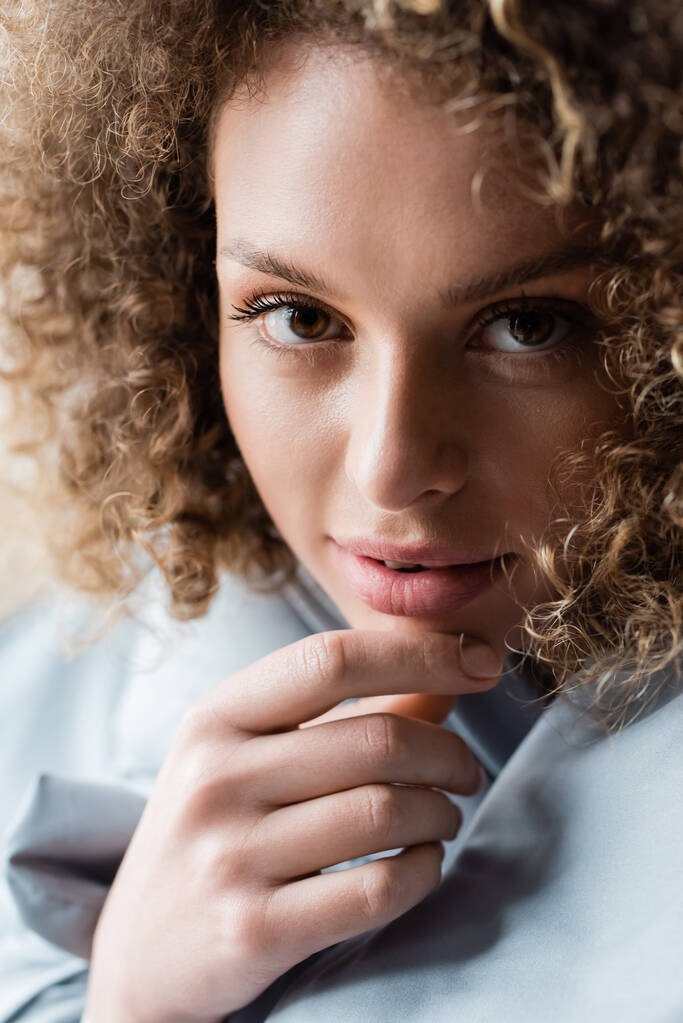 close up portrait of charming curly woman with brown eyes touching chin and looking at camera - Photo, Image