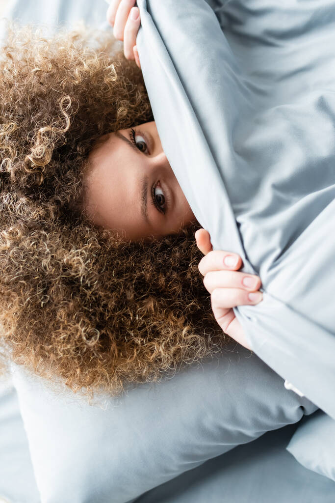 top view of curly woman obscuring face with blanket and looking at camera - Photo, Image