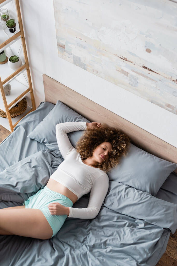 πάνω όψη της νεαρής γυναίκας με κλειστά μάτια ξαπλωμένη στο κρεβάτι με άσπρες πιτζάμες - Φωτογραφία, εικόνα