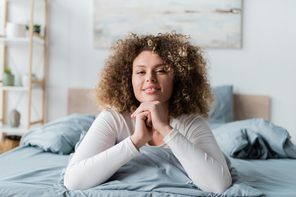 pleased curly woman smiling at camera while lying on bed - Photo, Image