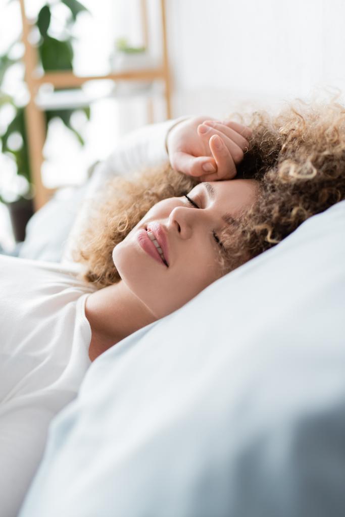 young curly woman smiling while sleeping on bed in morning - Photo, Image