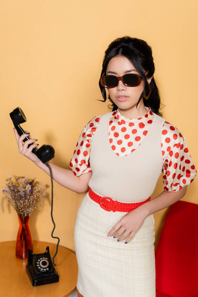 Stilvolle asiatische Frau mit Sonnenbrille und Hörer auf orangefarbenem Hintergrund - Foto, Bild