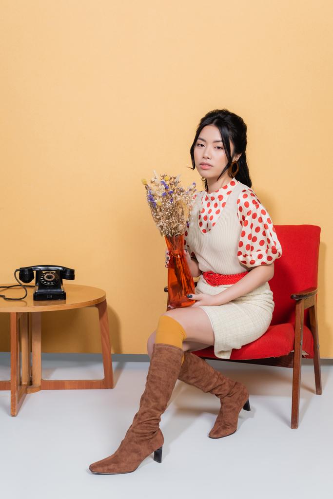 jong aziatisch model in vintage kleding houden bloemen terwijl zitten op fauteuil op oranje achtergrond - Foto, afbeelding