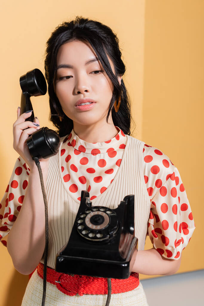 Стильная азиатская женщина держит ретро телефон на оранжевом фоне - Фото, изображение