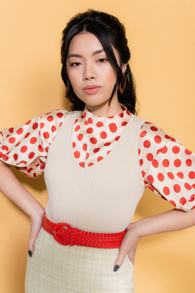 Portret stylowej azjatyckiej modelki trzymającej się za ręce na biodrach na pomarańczowym tle - Zdjęcie, obraz