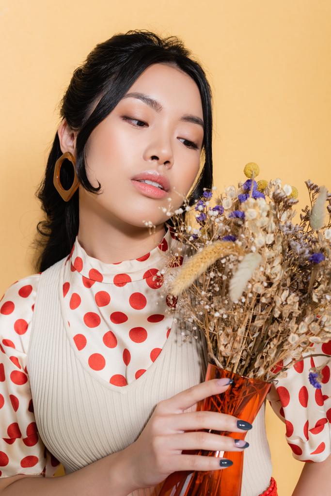 Portrait of stylish asian woman in retro clothes looking at flowers isolated on orange  - Photo, Image