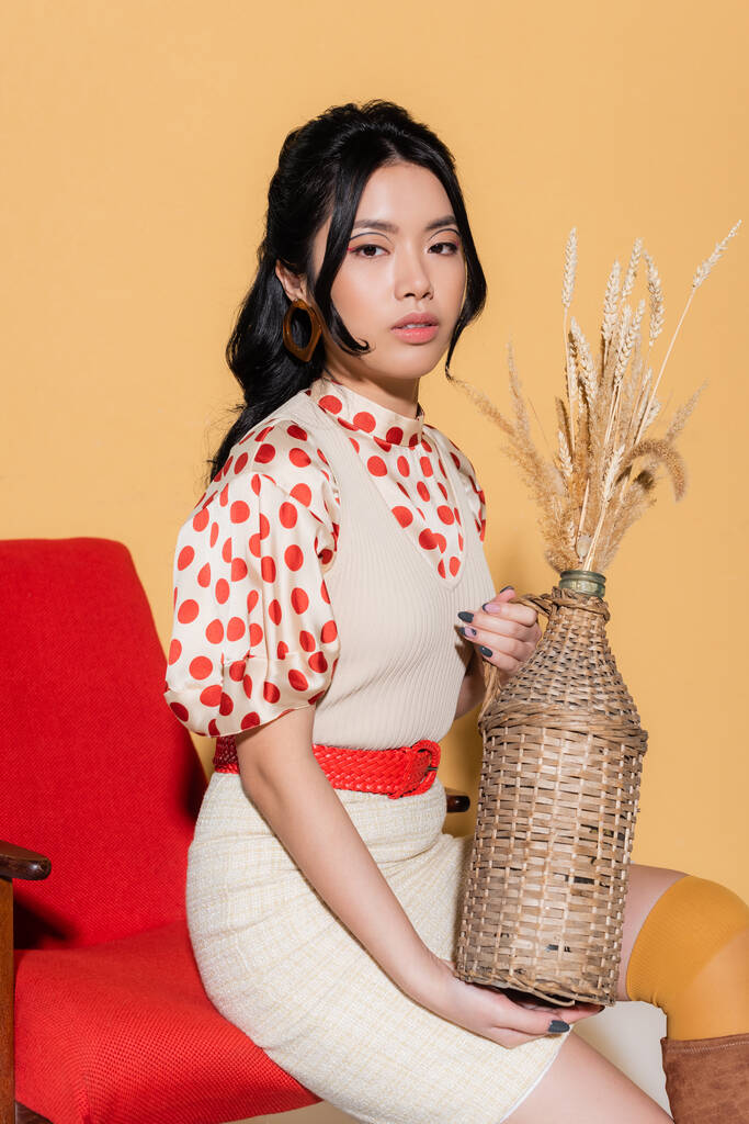 Trendy asian woman in blouse and dress holding vase with spikelets while sitting on armchair on orange background - Photo, Image