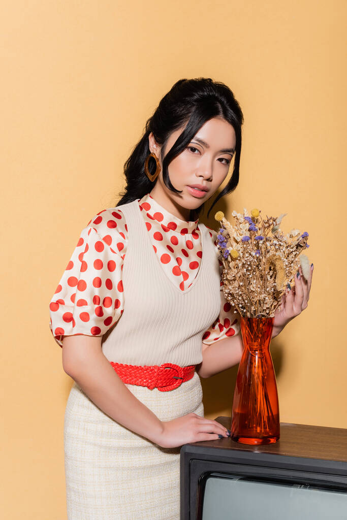 Πορτρέτο του κομψού ασιατικού μοντέλου σε vintage ρούχα αγγίζοντας τα λουλούδια στην τηλεόραση σε πορτοκαλί φόντο - Φωτογραφία, εικόνα