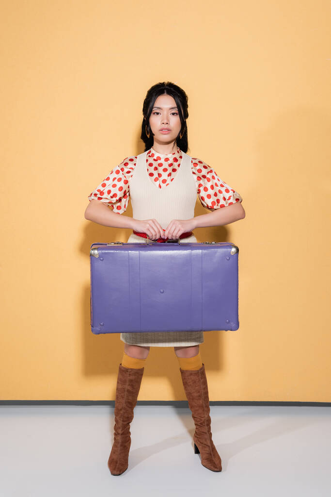 Longitud completa de elegante modelo asiático sosteniendo maleta retro sobre fondo naranja - Foto, imagen