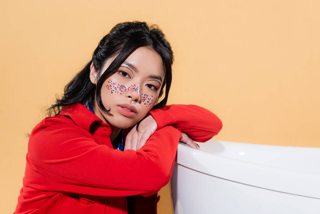 Asiatisches Modell mit Glitzern im Gesicht blickt in die Kamera in der Nähe der Badewanne isoliert auf orange  - Foto, Bild