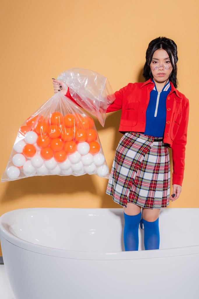 Joven modelo asiático en ropa vintage sosteniendo paquete con bolas en bañera sobre fondo naranja - Foto, imagen