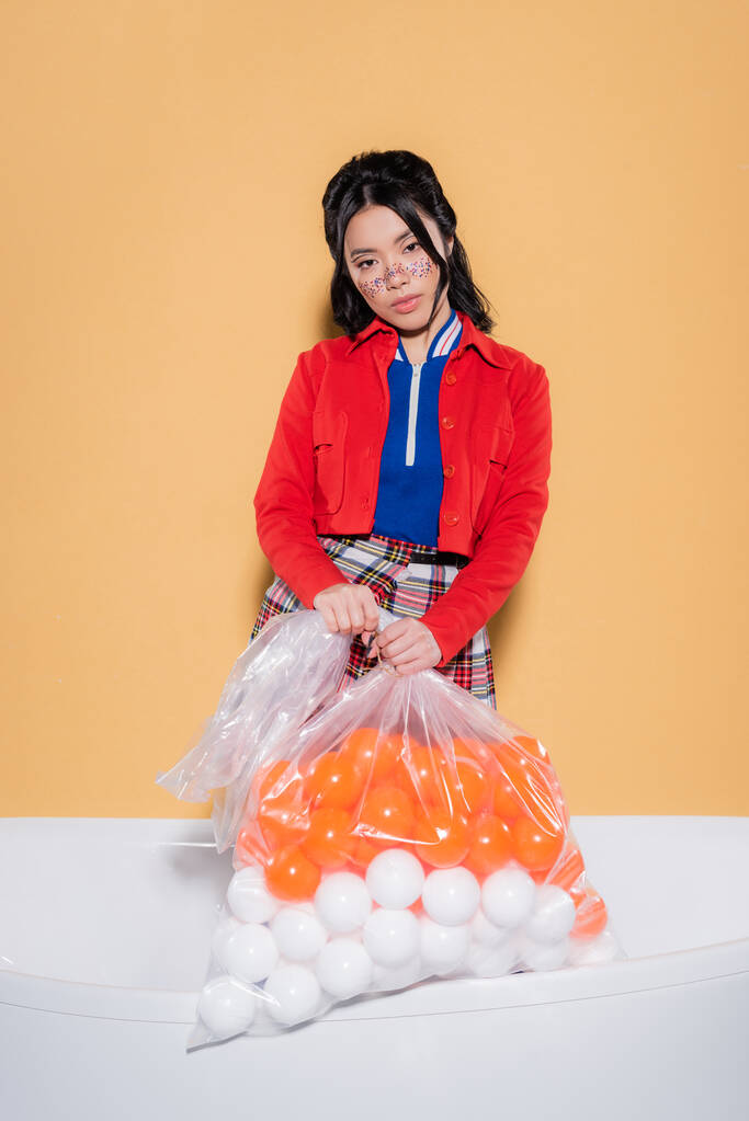 Mujer asiática de moda con maquillaje de purpurina sosteniendo bolsa de plástico con bolas en bañera sobre fondo naranja - Foto, Imagen