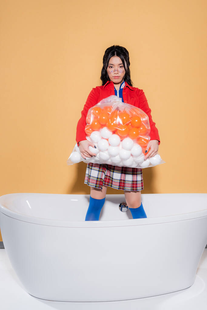 Elegante donna asiatica in possesso di sacchetto di plastica con le palle e guardando la fotocamera nella vasca da bagno su sfondo arancione - Foto, immagini