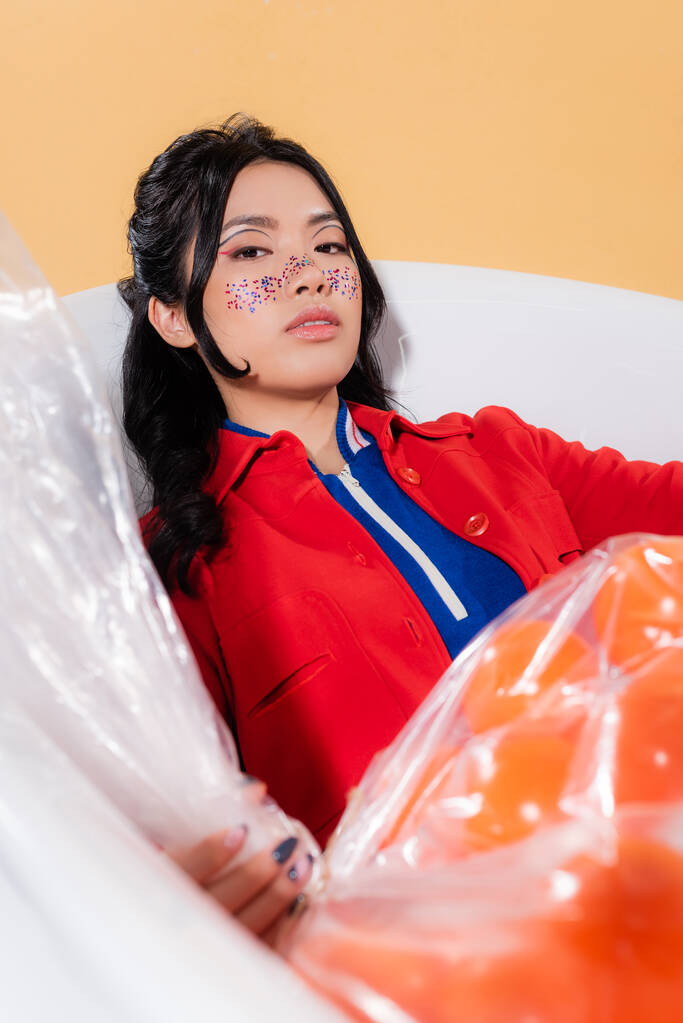 Retrato de mulher asiática elegante com maquiagem brilho posando no banho perto de saco de plástico borrado com bolas isoladas na laranja  - Foto, Imagem