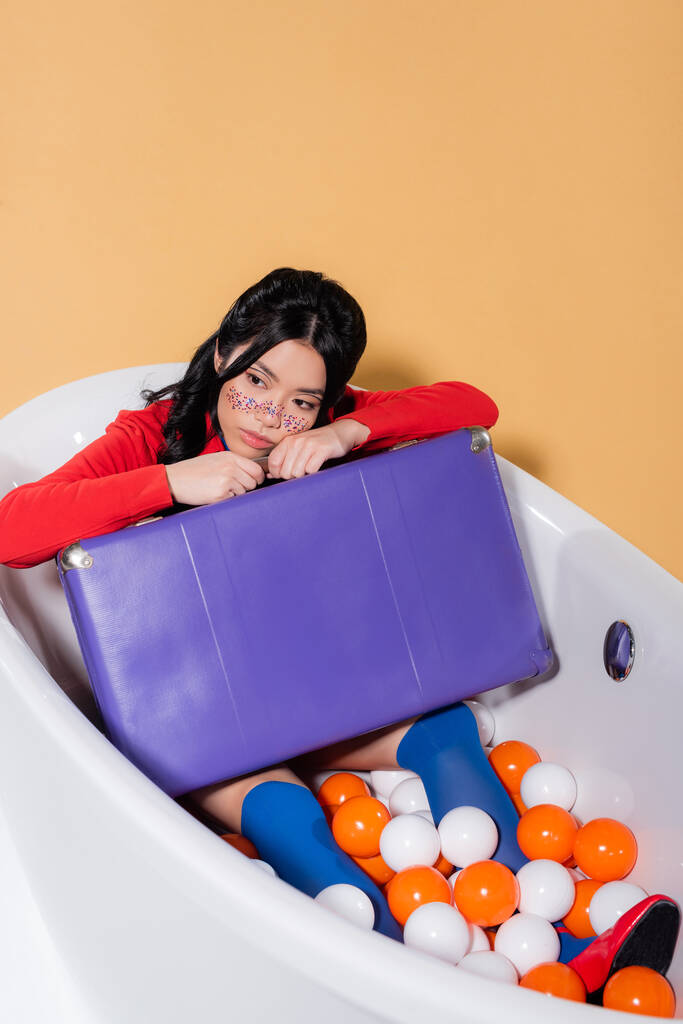Pěkný asijský model s třpytkami na obličeji drží retro kufr ve vaně s kuličkami na oranžovém pozadí - Fotografie, Obrázek