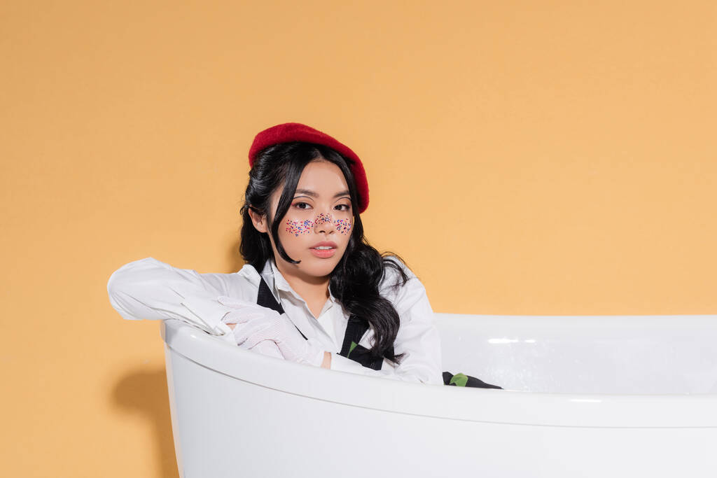 Elegante mujer asiática en guantes y boina sentada en bañera sobre fondo naranja - Foto, Imagen