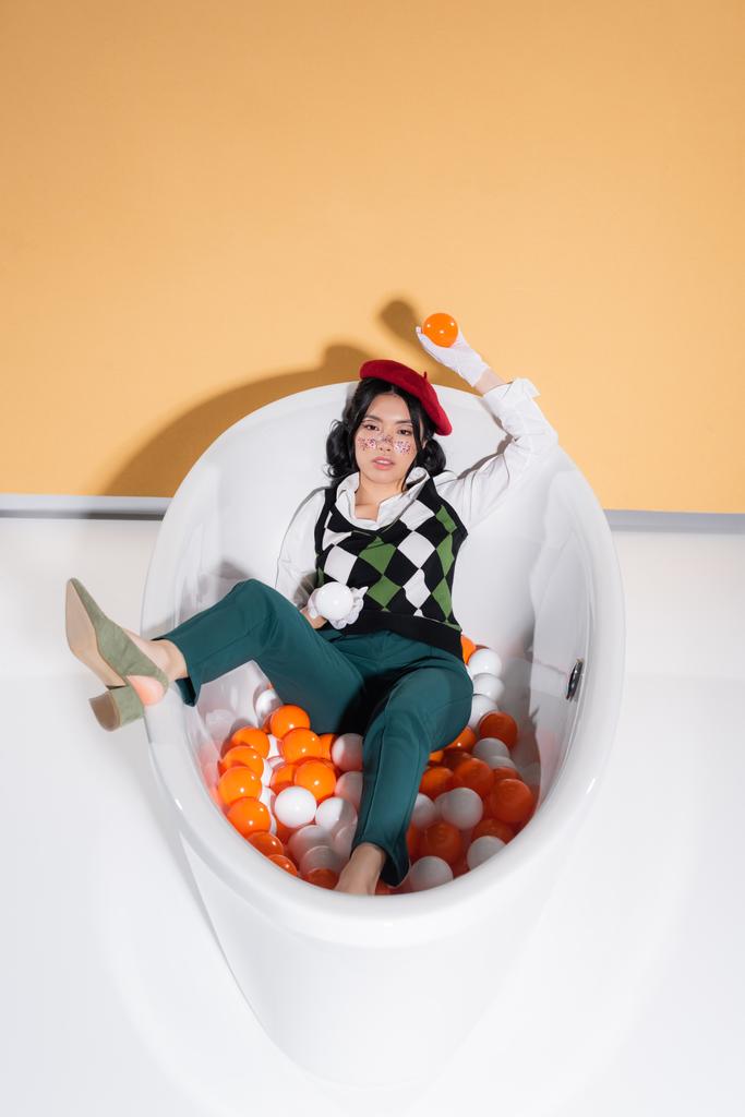 Trendy modello asiatico in berretto tenendo le palle e guardando la fotocamera nella vasca da bagno su sfondo arancione - Foto, immagini