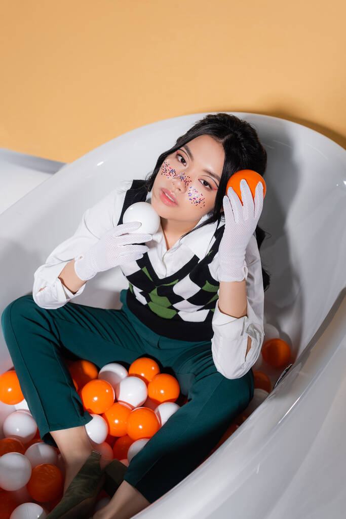 Alto ângulo vista da moda asiática modelo em luvas segurando bolas enquanto sentado na banheira no fundo laranja - Foto, Imagem