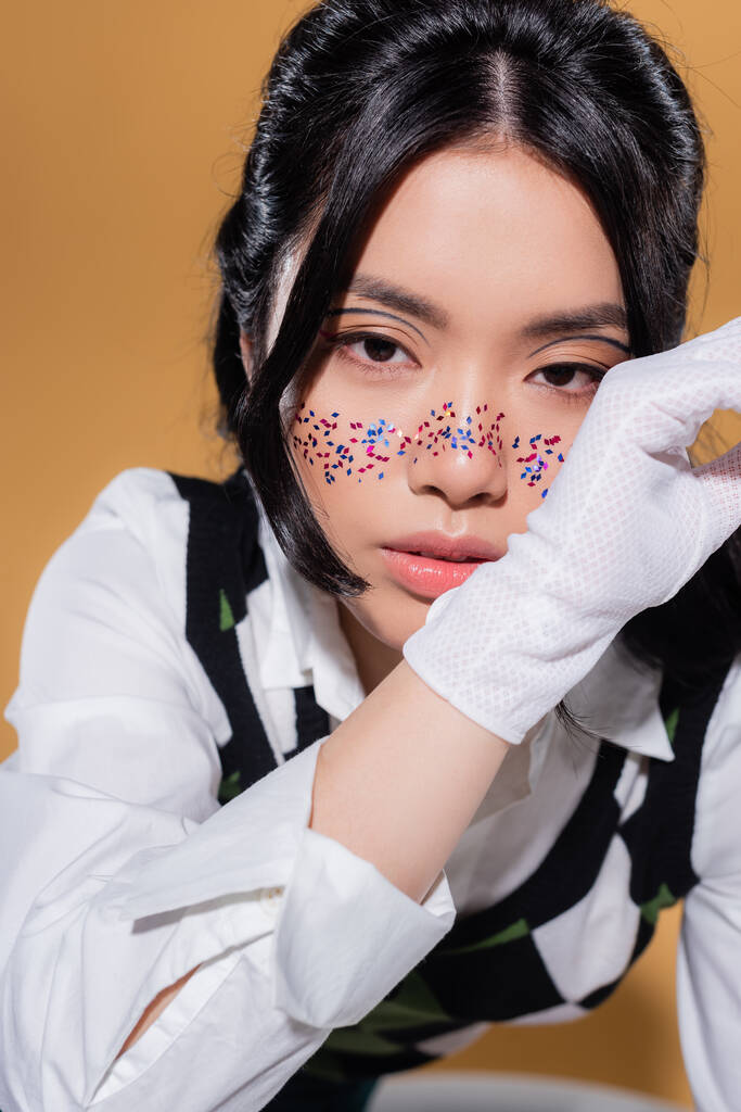 Portret van modieus aziatisch model met glitter make-up kijken naar camera op oranje achtergrond - Foto, afbeelding