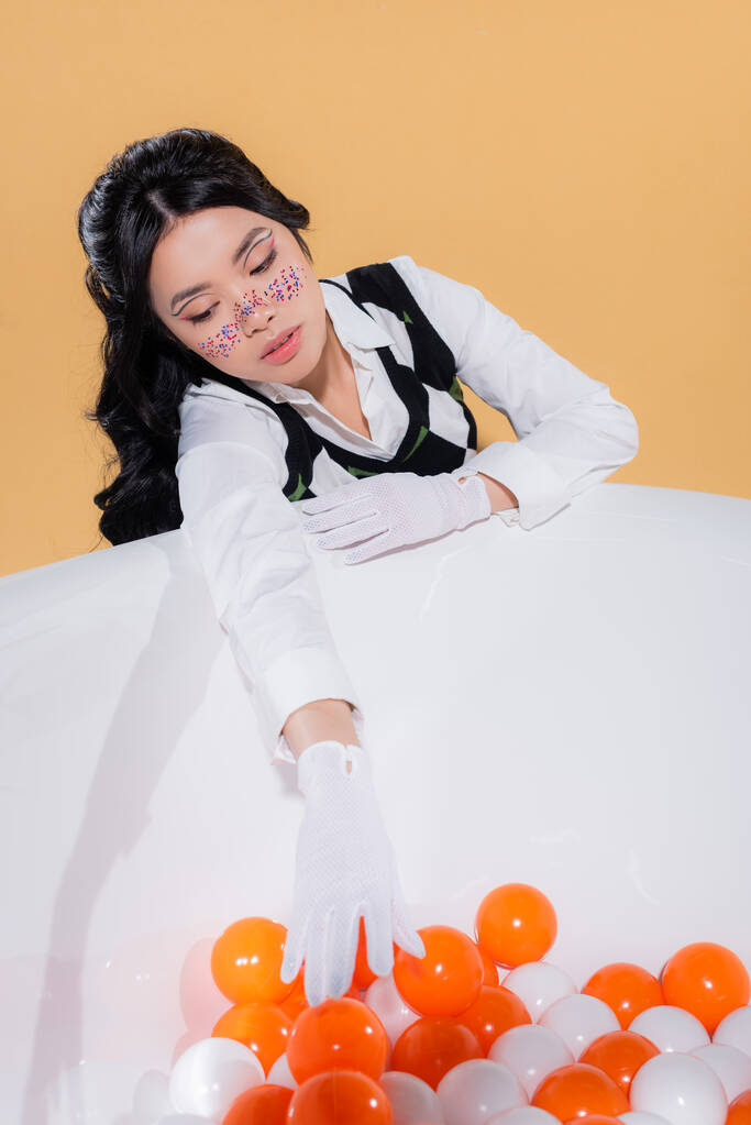 Fashionable asian model in gloves looking at balls in bathtub isolated on orange  - Photo, Image