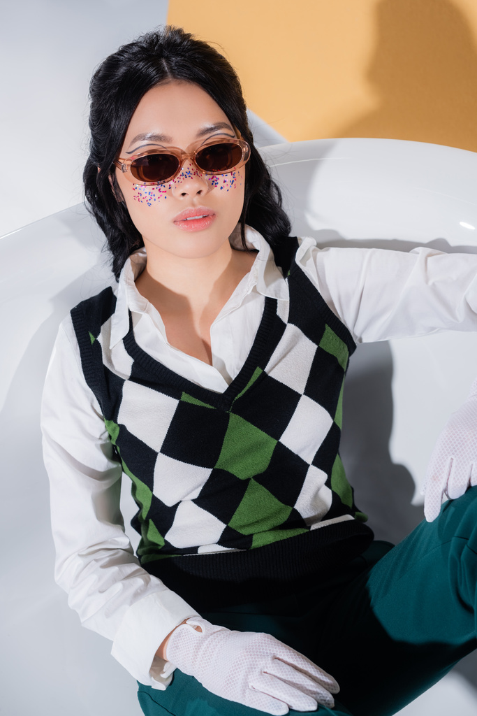 Azjatycki model w okularach przeciwsłonecznych i retro ubrania siedzi w wannie na pomarańczowym tle - Zdjęcie, obraz