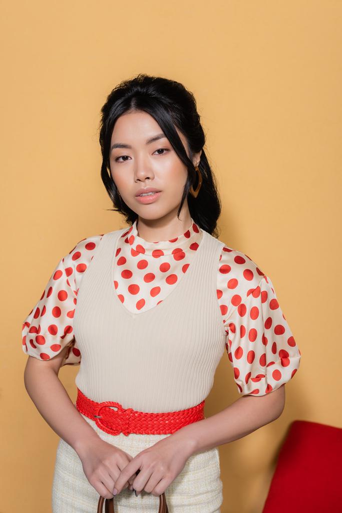 Joven modelo asiático en ropa vintage sosteniendo bolso de mano sobre fondo naranja - Foto, Imagen