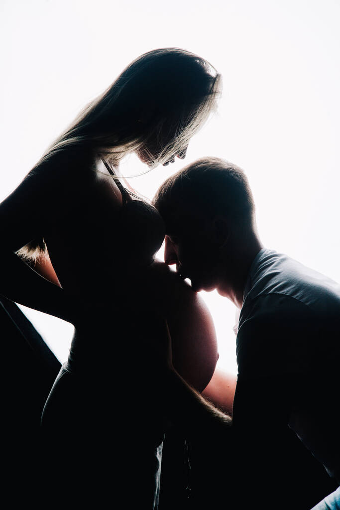 Муж целует беременную жену в живот. Темные силуэты на белом фоне. Фото из студии. Высокое качество фото - Фото, изображение