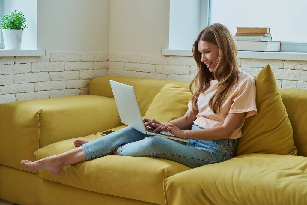 Приваблива молода жінка працює на ноутбуці і посміхається, сидячи на дивані вдома
 - Фото, зображення