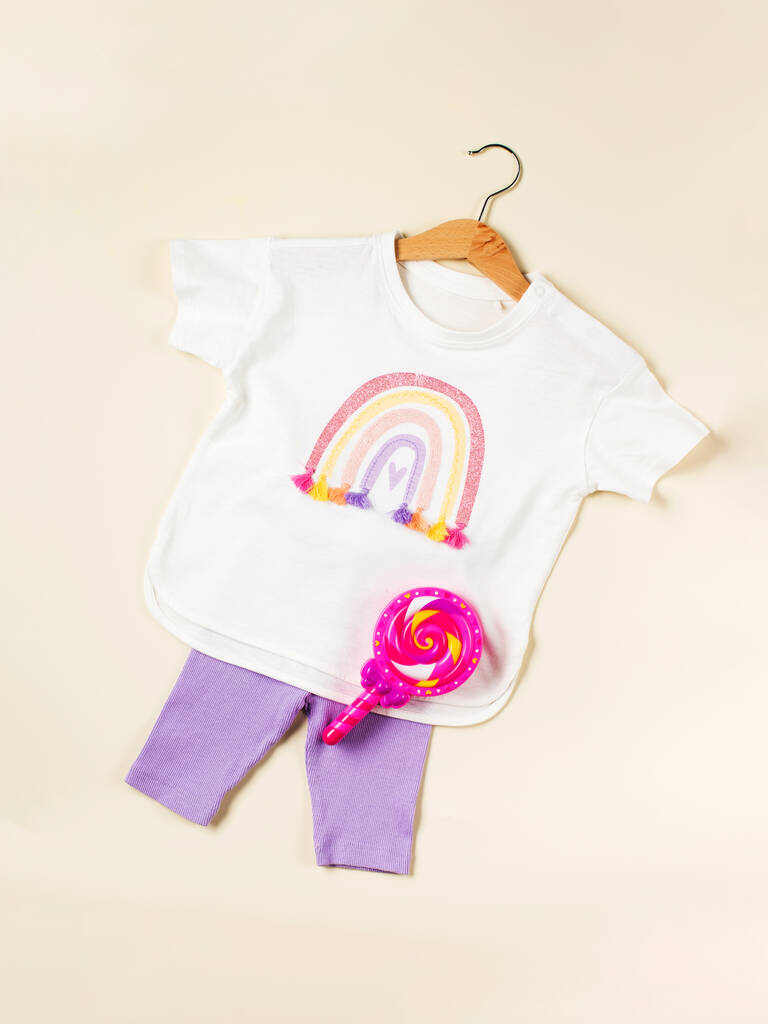 T-shirt pour enfant avec un arc-en-ciel sur un cintre et un short lilas sur fond beige. Vêtements pour enfants pour l'été. Pose plate. - Photo, image