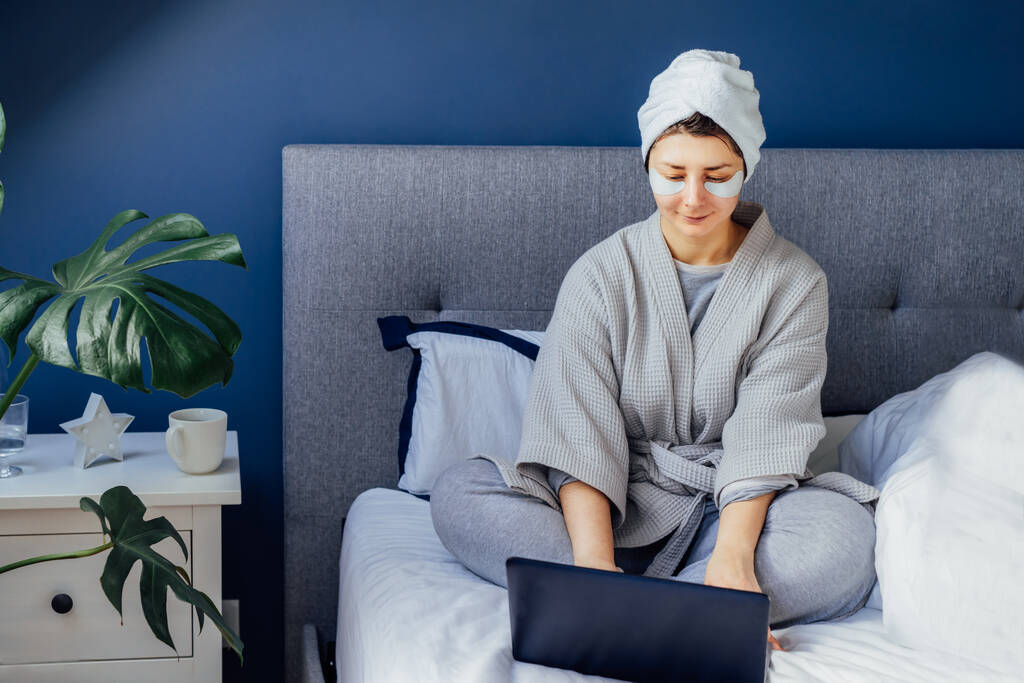 Vrouw in pyjama en badjas met oogvlekken en handdoek op haar hoofd met laptop terwijl ze op haar bed zat. Schoonheidsroutine tijdens het thuiswerken. Goedemorgen gewoonten. Werk vanuit huis. Selectieve focus. - Foto, afbeelding