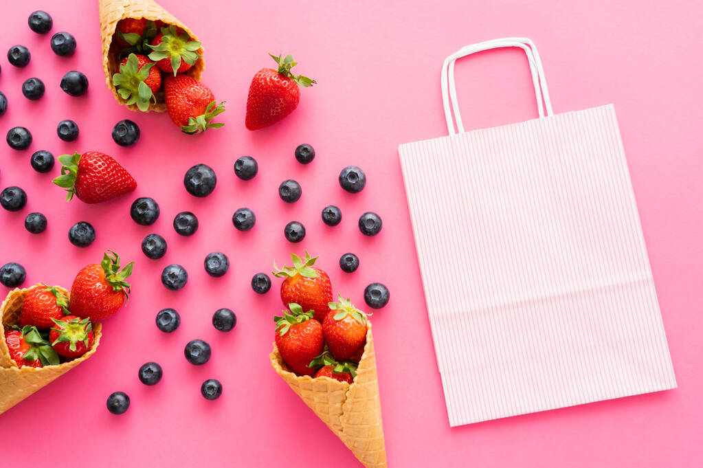 Piso con bolsa de compras cerca de bayas frescas y conos de waffle sobre fondo rosa  - Foto, imagen