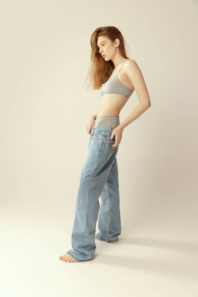 Portret van slanke mooie jonge vrouw poserend in grijs ondergoed en oversized jeans geïsoleerd over grijze studio achtergrond. Concept van schoonheid, gezondheid, lichaams- en huidverzorging, spa, cosmetologie, fitness, reclame - Foto, afbeelding