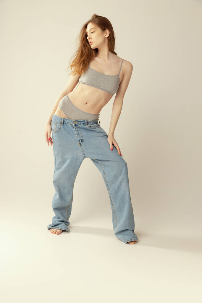Одна струнка красива молода дівчина позує в сірій нижній білизні і великі джинси ізольовані на сірому студійному фоні. Втрата ваги. Концепція краси, здоров'я, догляд за тілом і шкірою, спа, косметологія, фітнес, реклама
 - Фото, зображення