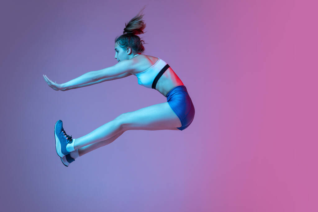 Momentos. Jovem, atleta feminina treinando técnica de salto em distância isolado no fundo do estúdio rosa com filtro de néon azul, luz. Conceito de ação, movimento, velocidade, estilo de vida saudável. Espaço de cópia - Foto, Imagem