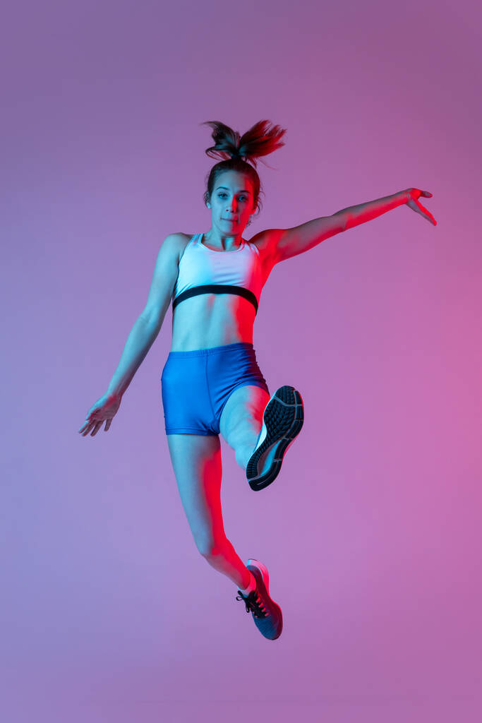 Training in long jump sport. Young girl, female athlete practicing isolated on pink studio background with blue neon filter, light. Concept of action, motion, speed, healthy lifestyle. Copy space - Photo, Image