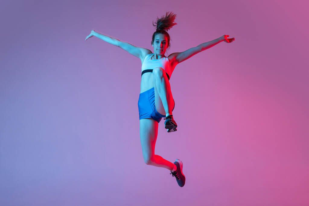 Anlar. Genç kız, uzun atlama tekniği eğitimi alan bayan atlet pembe stüdyo arka planında mavi neon filtresi ve ışık ile izole edilmiş. Eylem kavramı, hareket, hız, sağlıklı yaşam tarzı. Boşluğu kopyala - Fotoğraf, Görsel