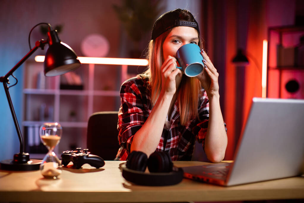 Αυτοπεποίθηση νεαρή γυναίκα πίνοντας καφέ ή τσάι, ενώ πληκτρολογείτε στο φορητό υπολογιστή στο γραφείο. Καυκάσια εργαζόμενη που κάθεται στο τραπέζι με ανοιχτό φορητό υπολογιστή, εργάζεται στο σύγχρονο γραφείο ή στο σπίτι το βράδυ. - Φωτογραφία, εικόνα