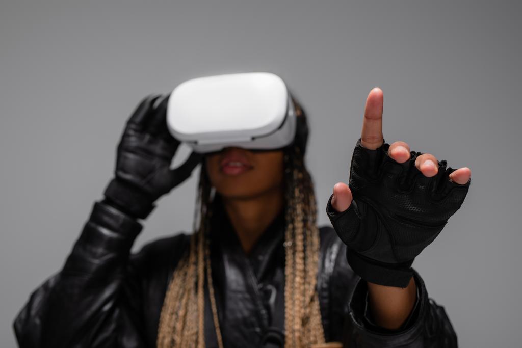 Unscharfe Afroamerikanerin mit Virtual-Reality-Headset und Handschuhen, die mit dem Finger auf grau zeigen  - Foto, Bild