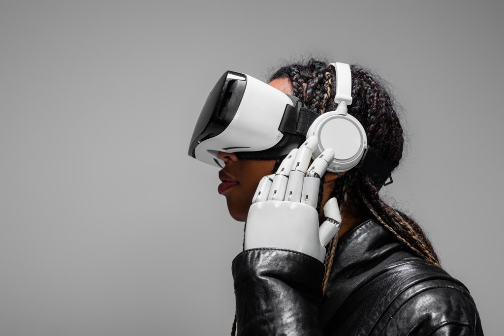 Zijaanzicht van Afro-Amerikaanse vrouw met robothand en vr headset luisteren muziek in koptelefoon geïsoleerd op grijs  - Foto, afbeelding
