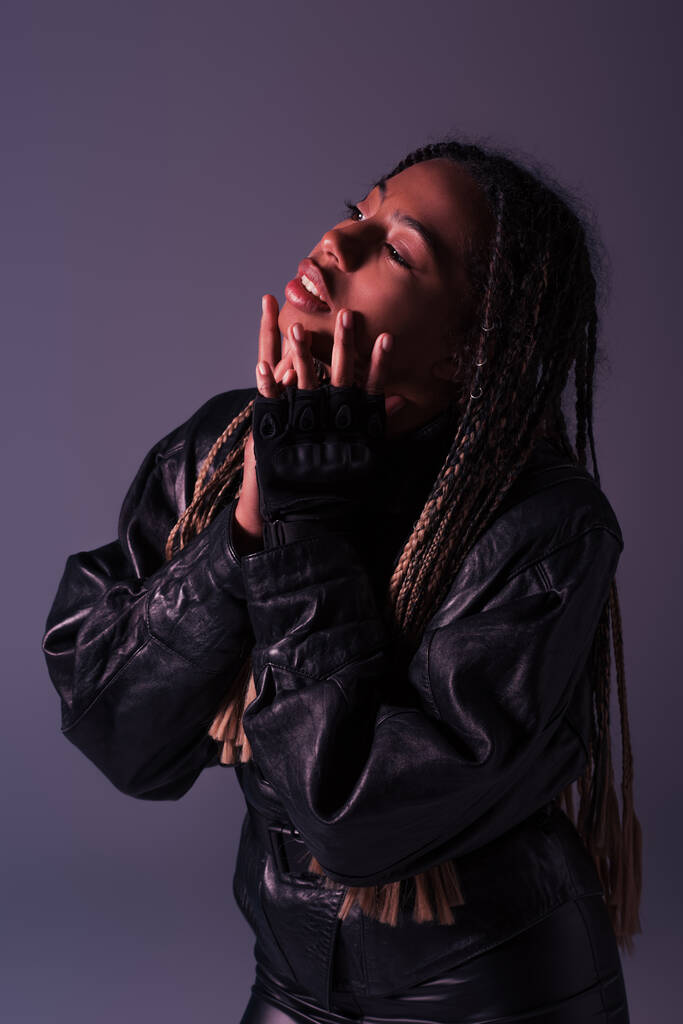 African american woman in leather jacket and glove touching face isolated on purple  - Photo, Image