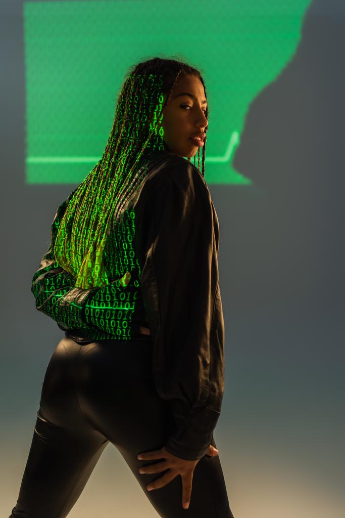 Mujer afroamericana con estilo con proyección de código informático en la chaqueta de cuero sobre fondo gris  - Foto, Imagen