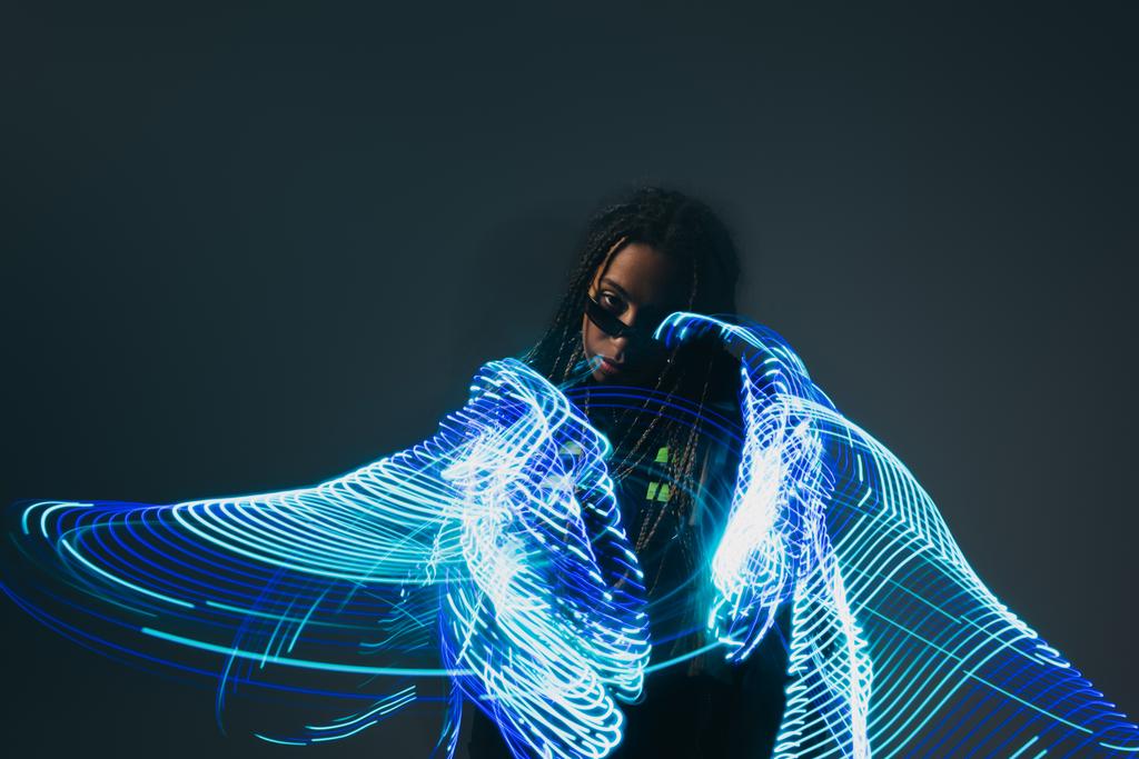 African american woman in smart glasses looking at camera near abstraction neon lights on grey background  - Photo, Image