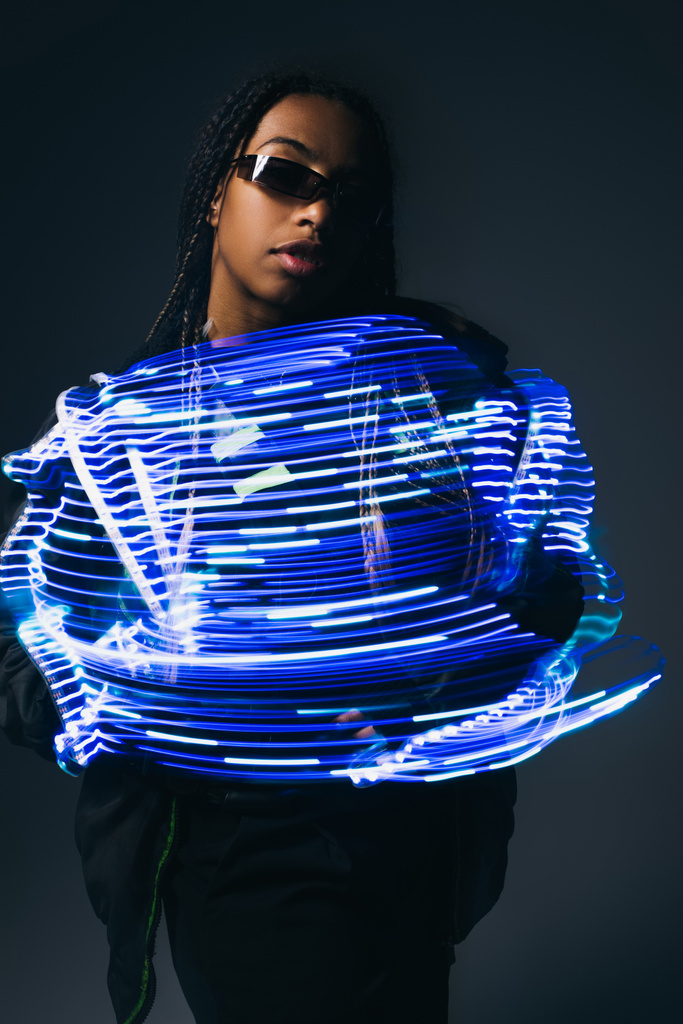 Stylish african american woman in smart glasses posing near neon lines on grey background  - Photo, Image