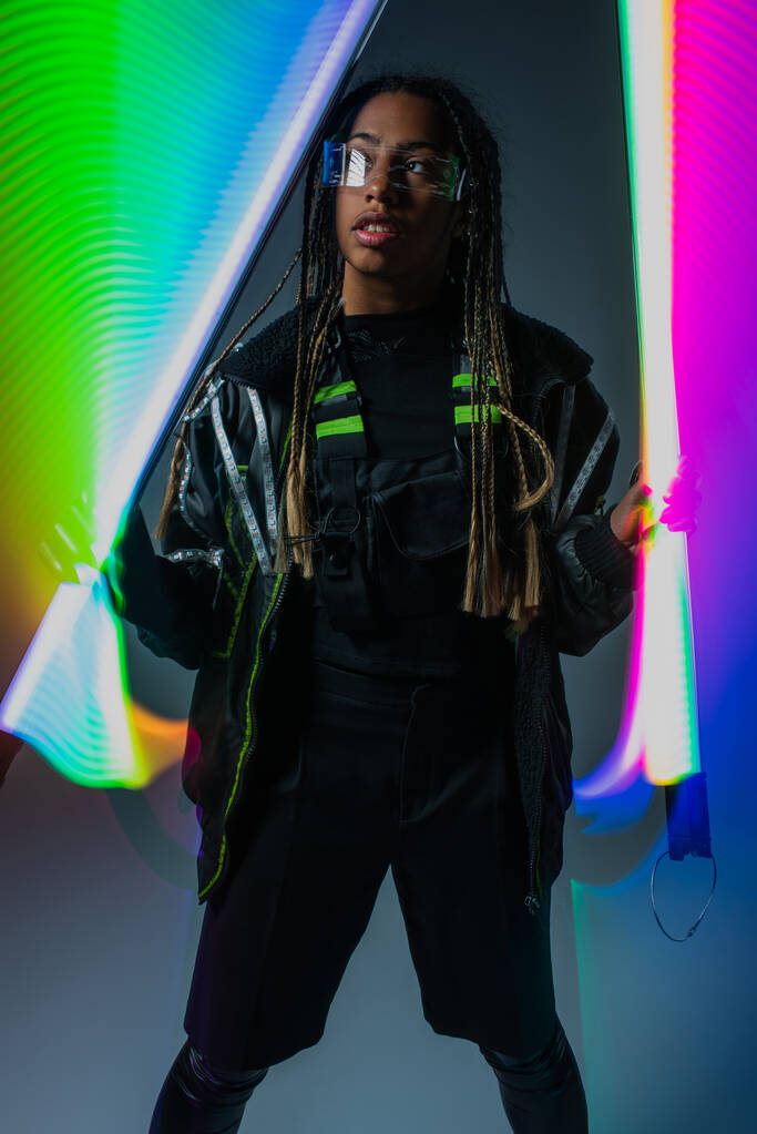 Stylish african american woman in smart glasses posing with neon sticks on grey background  - Photo, Image