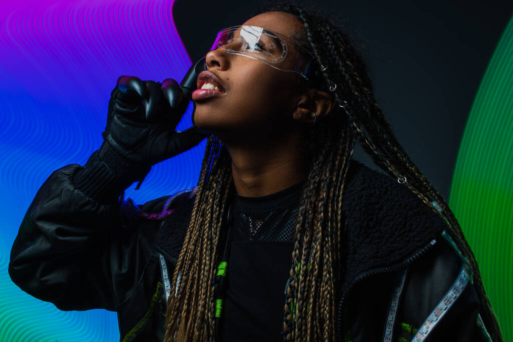 Африканская американка в перчатках и умных очках, смотрящая в сторону яркой абстракции на сером фоне  - Фото, изображение