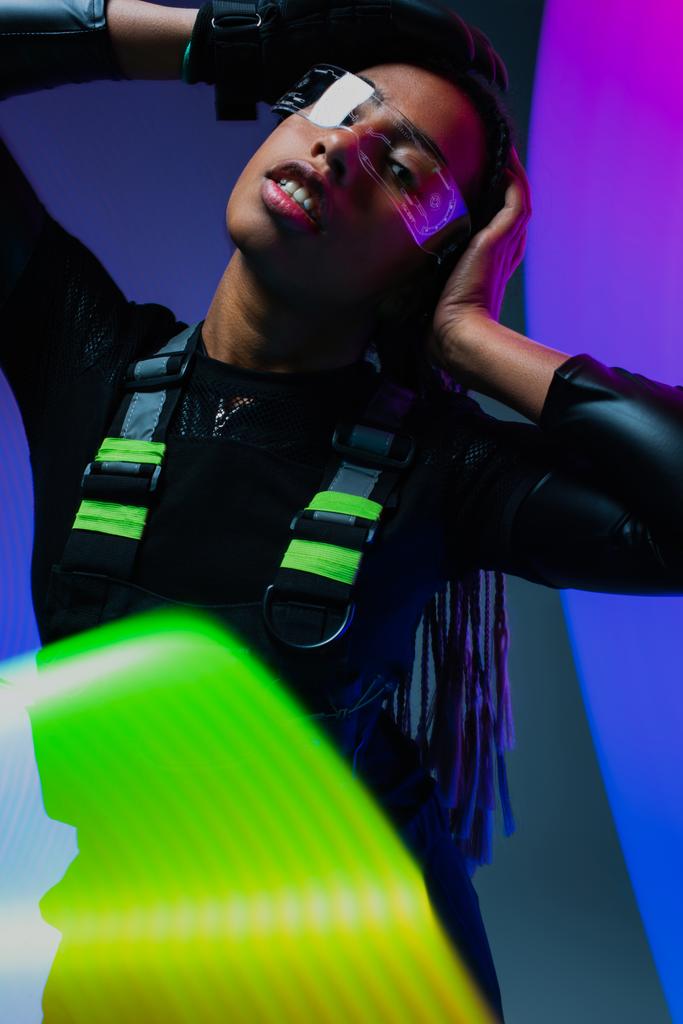 Стильна афро-американська жінка в розумних окулярах, що виставляється біля неонових вогнів на барвистому тлі  - Фото, зображення